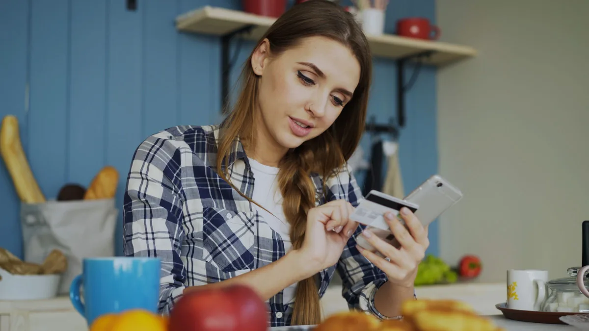 A woman checking her phone and a credit card while shopping online from her kitchen
